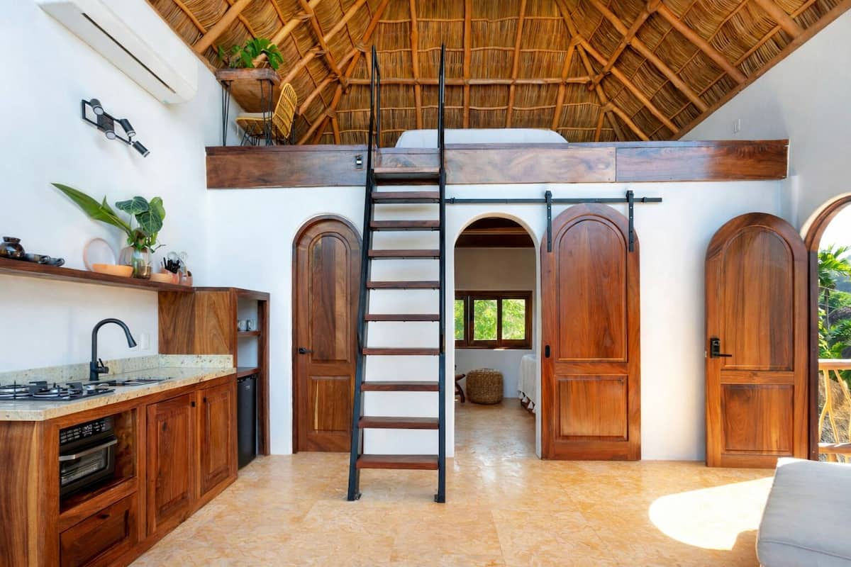 Una cocina con una isla central, armarios de madera, horno empotrado y ventilador de techo. Hay dos puertas de madera y una ventana con vistas a zonas verdes.