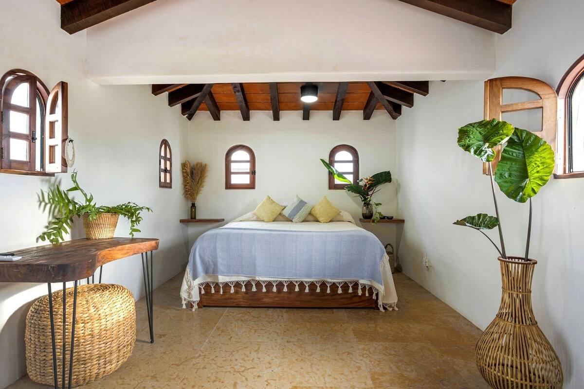 Un dormitorio con una cama, una mesa de madera, un reposapiés de mimbre y una planta grande.