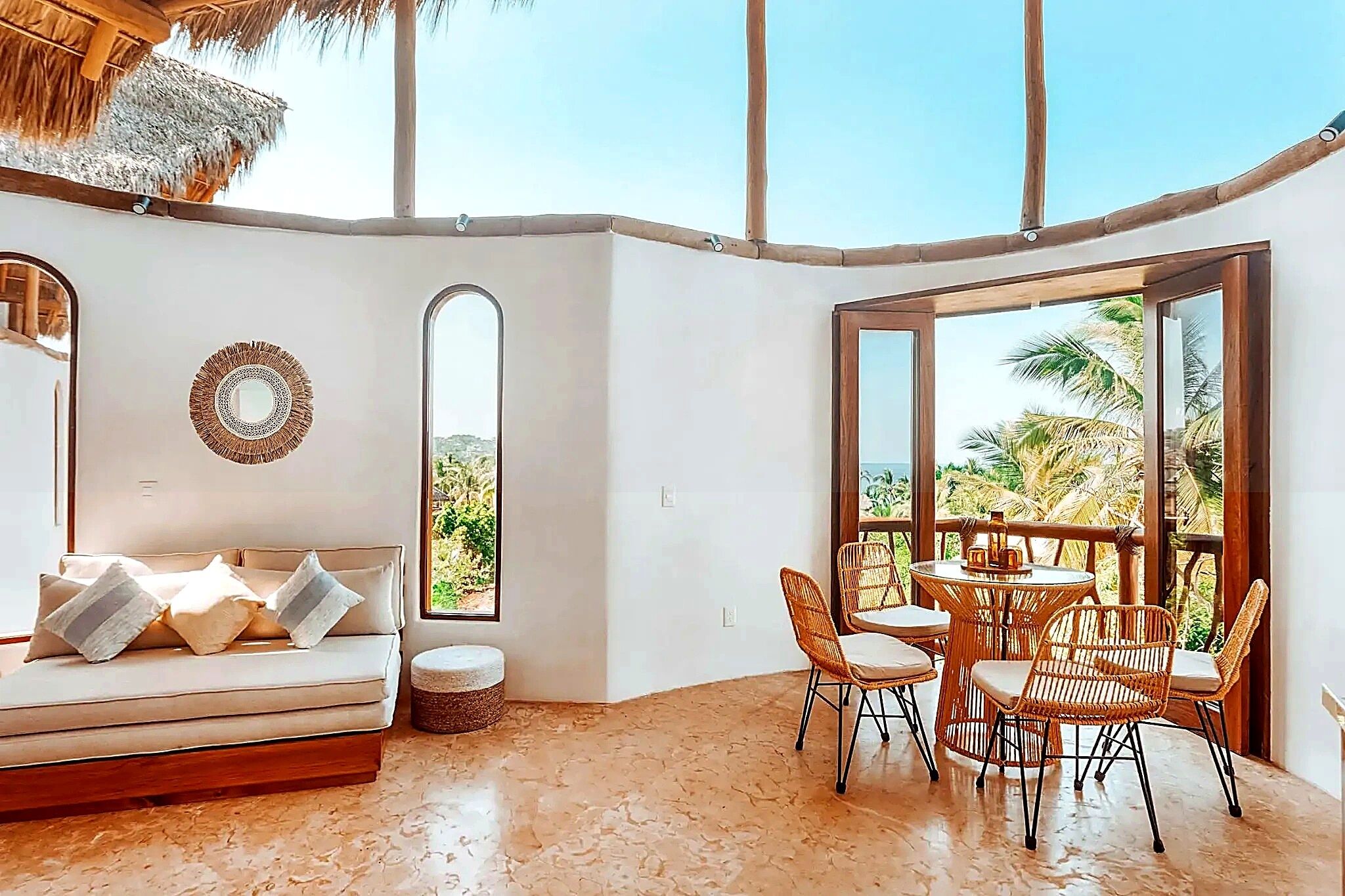 A room with a sofa, dining table, chairs, and a large window with a view of palm trees.