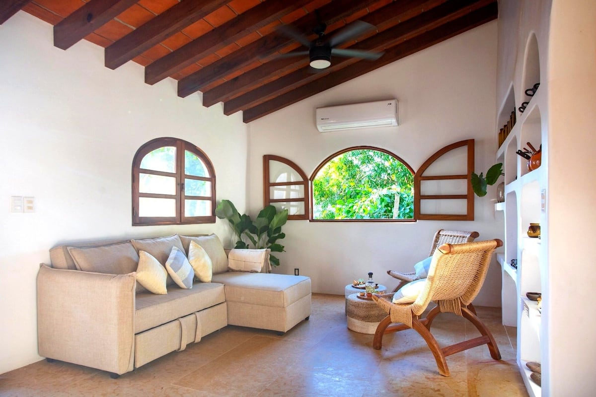 Una sala de estar con un sofá, una silla y una pequeña mesa. Se observan ventanas de arco y un ventilador de techo.