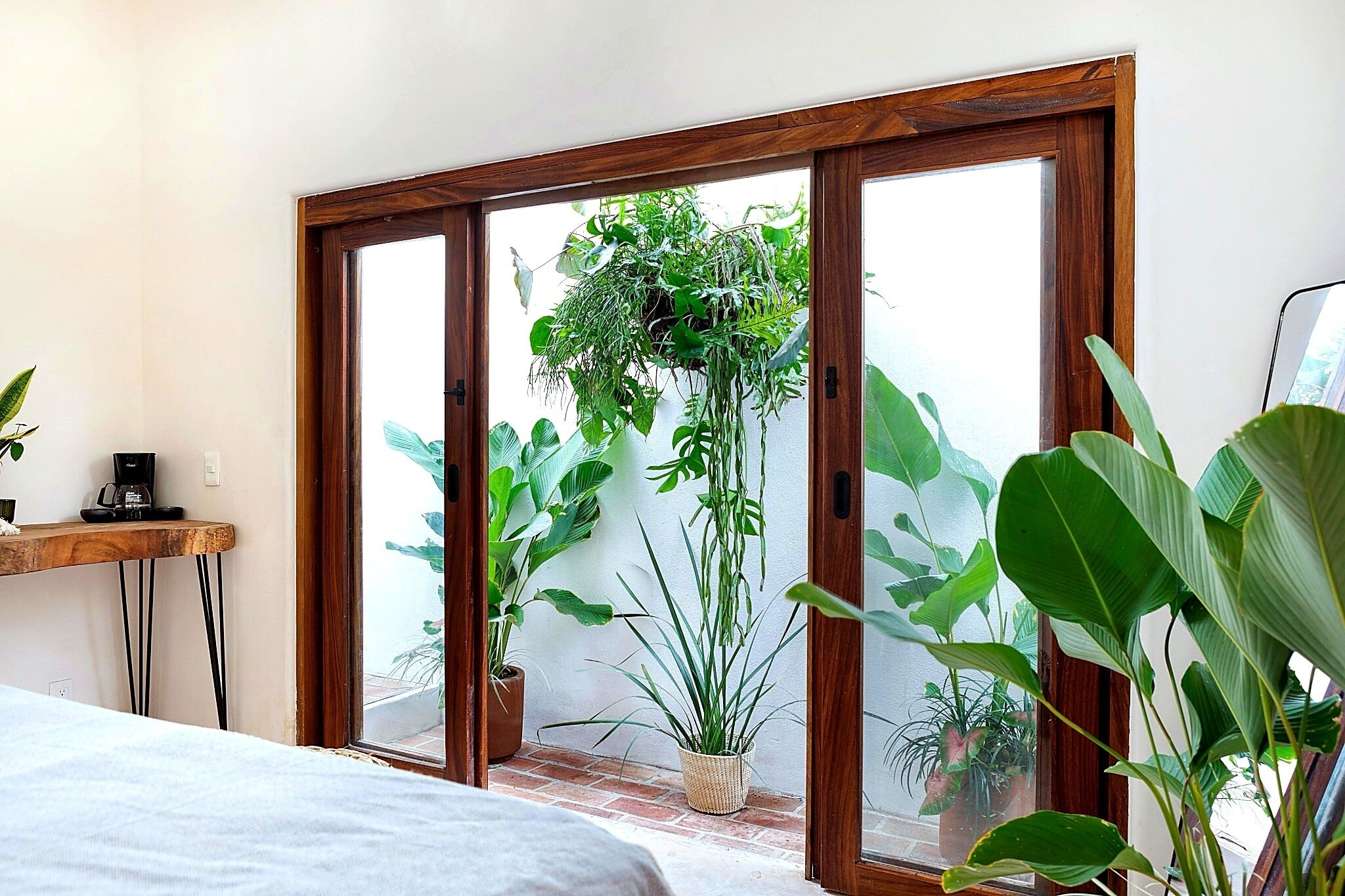 Una habitación con cama, una mesa de madera con cafetera y un amplio ventanal corredero que da a un balcón con plantas en macetas.