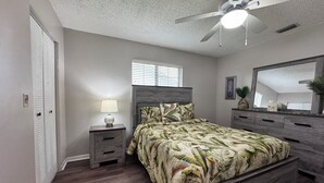 Une chambre à coucher comprenant un lit, une table de chevet, une commode, un ventilateur de plafond et une fenêtre avec des stores.