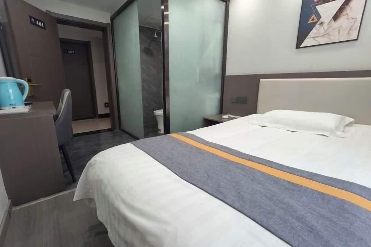 A hotel room with a bed, a desk, a chair, a coffee maker, and a shower area.