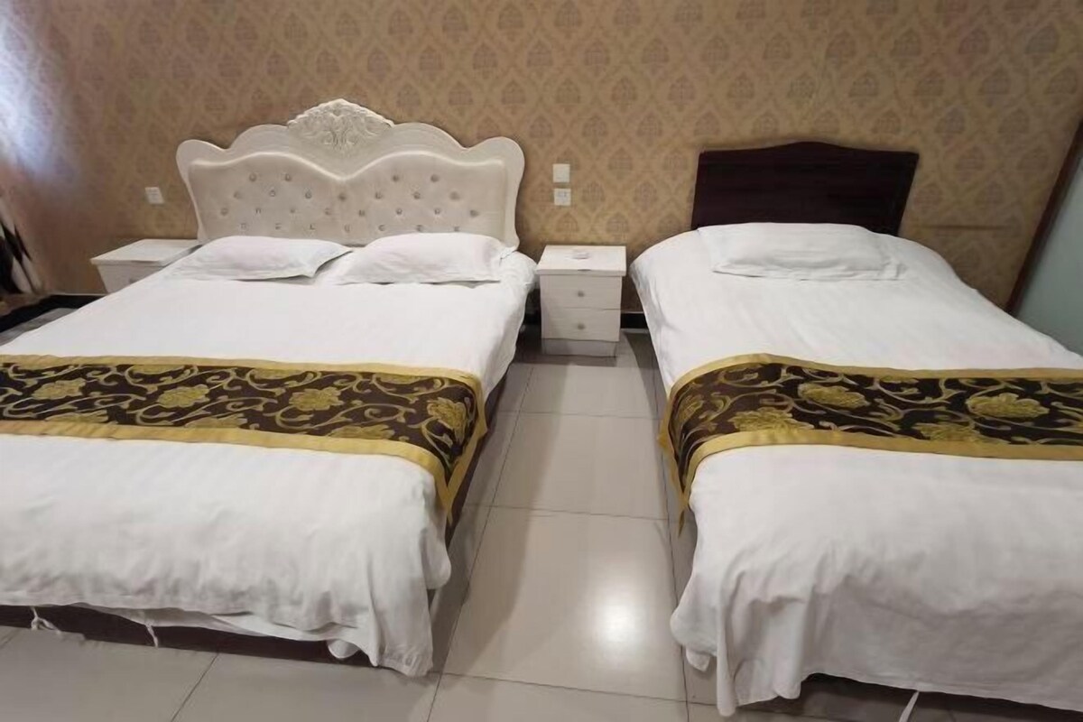 A hotel room with two beds, each with a white headboard and a patterned bedspread. There are bedside tables with lamps and a small drawer.