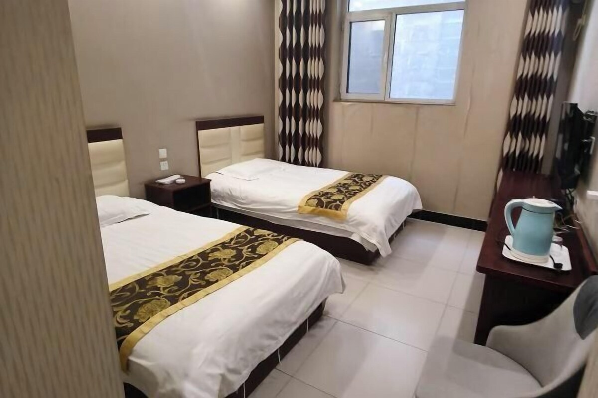 A hotel room with two beds, a desk with a kettle, and a window with curtains.