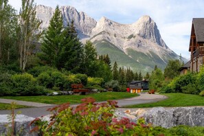 A mountain lodge with a stone exterior, surrounded by lush greenery and a prominent mountain in the background.
