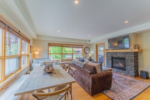 A spacious living area with a fireplace, a television mounted above it, and a large window providing a view of the outdoors. - Luxury Corner Condo w/ Mountain Views + Hot Tub (Canmore)