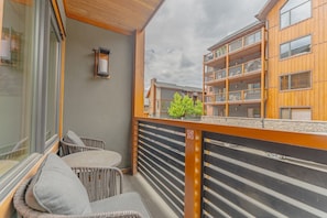 A balcony with wicker furniture, a stone wall, and a view of a multi-story building. - Luxury Corner Condo w/ Mountain Views + Hot Tub (Canmore)