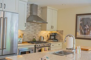 A modern kitchen with stainless steel appliances, a brick backsplash, and a coffee maker. - Luxury Corner Condo w/ Mountain Views + Hot Tub (Canmore)