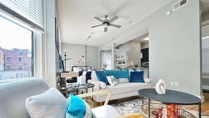 A modern living room with a gray sofa, a wooden chair, and a round coffee table. There is a ceiling fan and a window with blinds. - Boho Studio Loft in OTR Near Downtown Cincy (Cincinnati)