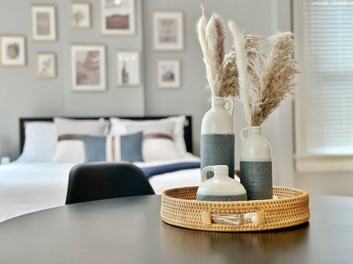 Une chambre d’hôtel avec un lit parfaitement fait, une table à manger agrémentée d’un set de table en fibres tressées, et deux vases contenant des plumes d’herbe de la pampa.