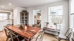Un vaste espace de salle à manger avec une table en bois et des chaises en métal, une cuisine à l’arrière-plan et une grande fenêtre avec des stores.
