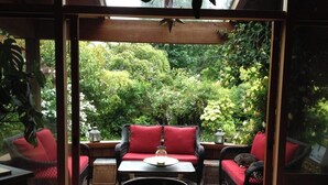 An outdoor seating area with wicker furniture and red cushions, a table with a lantern, and a view of lush greenery. - Roberts Creek: Charming 3-bedroom/2 bath post 'n beam steps to Henderson Beach (Roberts Creek)