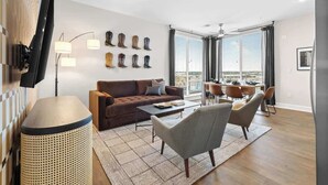 A modern living room with a brown sofa, a coffee table, and a TV mounted on the wall. There are multiple windows with blinds, a dining area with chairs, and a decorative wall with boots. - 2 King Suites Walk to Broadway & Gulch (Nashville)
