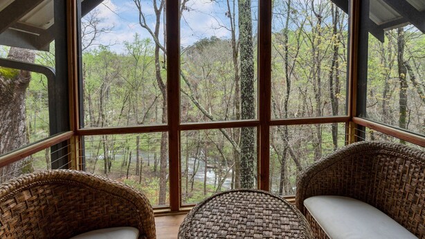 A room with large windows offering a view of a forest. There are two wicker chairs with cushions. - Cozy, creekside cottage in beautiful Dawsonville (Dawsonville)