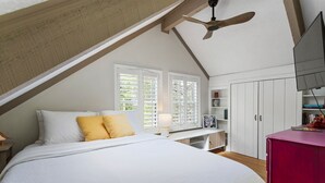 A bedroom with a sloped ceiling, a bed with white bedding and a yellow pillow, a desk with a lamp, a pink cabinet, and a TV on the wall. - Cozy, creekside cottage in beautiful Dawsonville (Dawsonville)