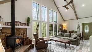 A cozy living room with a stone fireplace, a comfortable sofa, and a large window offering a view of trees. - Cozy, creekside cottage in beautiful Dawsonville (Dawsonville)