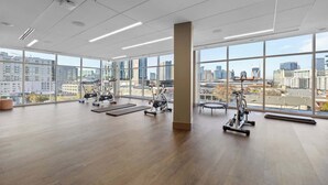 Un gimnasio espacioso con amplias ventanas que ofrecen vistas de la ciudad, equipado con diversas máquinas de ejercicio y colchonetas.