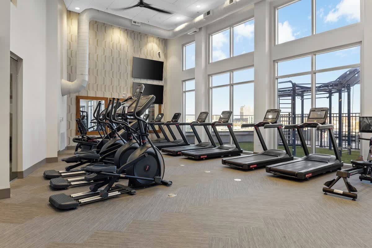 Un gimnasio con cintas de correr y máquinas elípticas dispuestas en filas, ventanas amplias y un ventilador de techo.