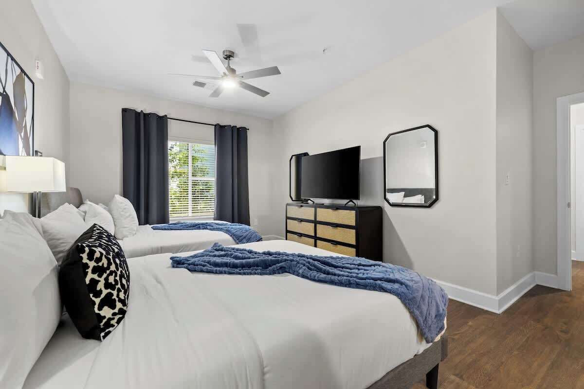 A hotel room with two beds, a ceiling fan, a TV, a mirror, and a window with curtains.