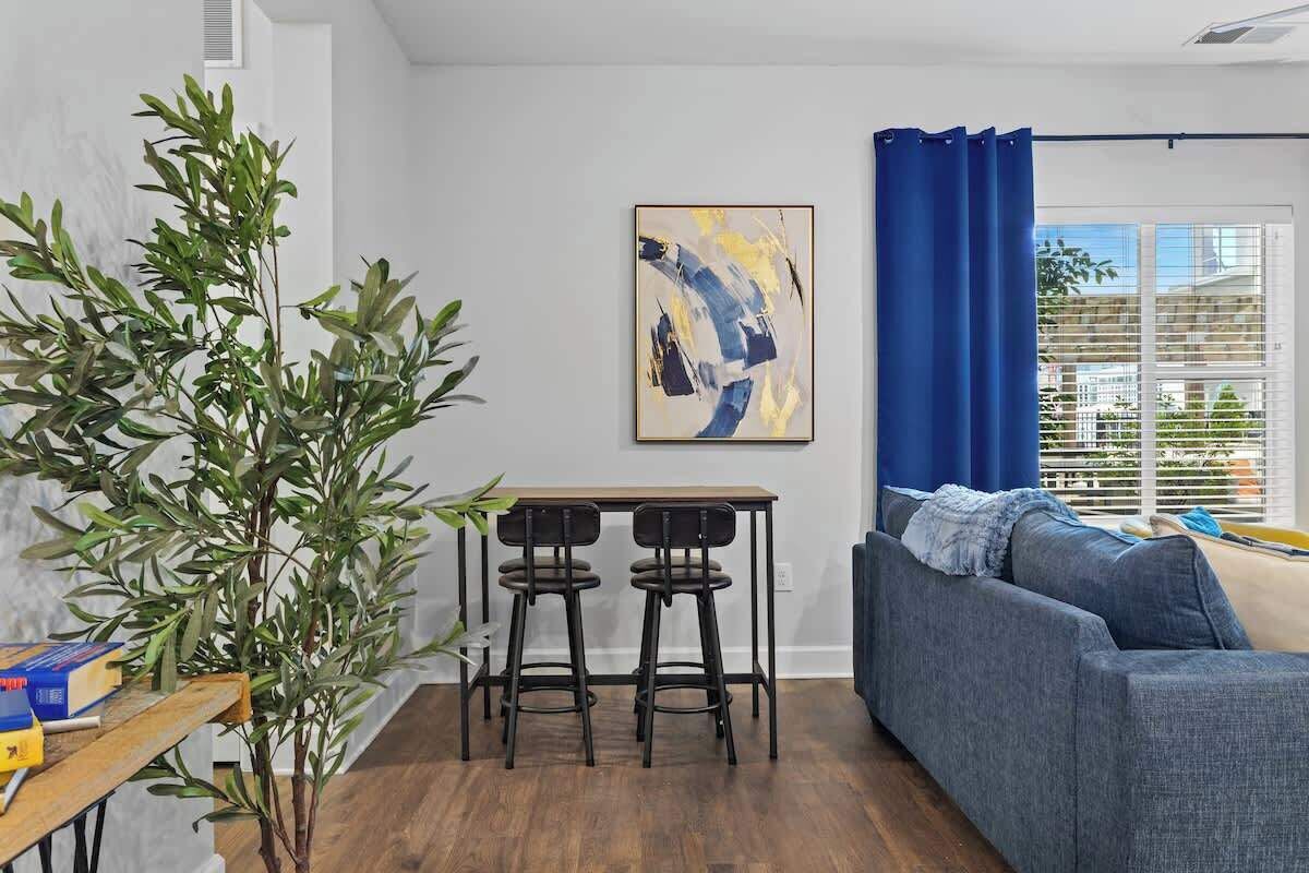 Una sala de estar con un sofá azul, una mesa de madera con dos sillas, un cuadro abstracto grande en la pared y una ventana con cortinas azules.