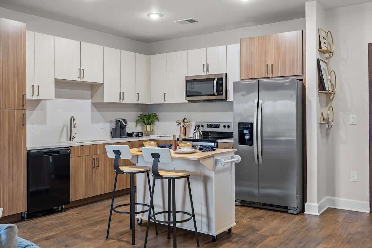 A modern kitchen with white cabinets, a stainless steel refrigerator, a microwave, and a coffee machine. There are two bar stools and a small island with a sink.