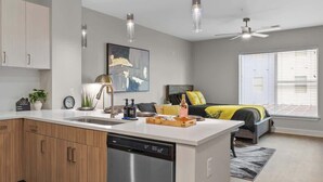 A modern kitchen with a dishwasher, a dining area with a table and chairs, a bed with yellow pillows, and a large window with blinds. - Luxe Gulch 1BR near Broadway | Sleeps 6 (Nashville)