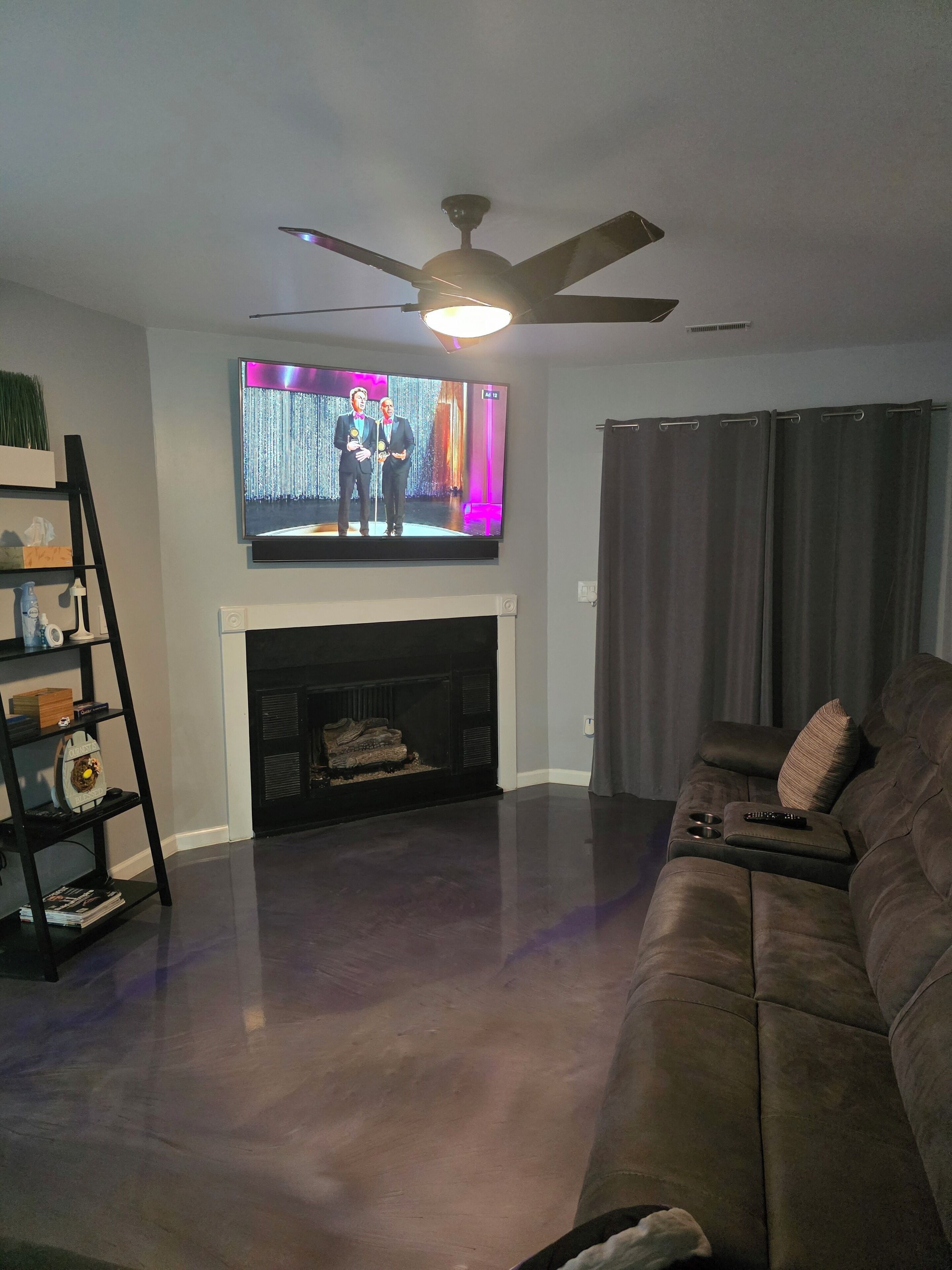 A living room with a fireplace, a television mounted above it, a ceiling fan, and a dark-colored couch.
