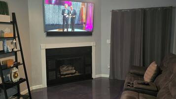 A living room with a fireplace, a television mounted above it, a ceiling fan, and a dark-colored couch.