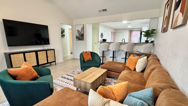 A spacious living room with a large sectional sofa, a green armchair, and a coffee table. A flat-screen TV is mounted on the wall, and there are framed pictures on the wall. - Swim, Hike, Explore & Relax just minutes from lakes-beaches, nature & Ft. Worth! (Azle)