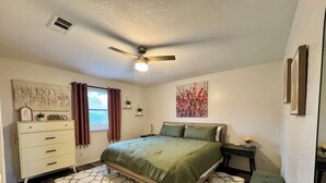 A bedroom with a bed, a dresser, a nightstand, a bench, a ceiling fan, and artwork on the walls. - Swim, Hike, Explore & Relax just minutes from lakes-beaches, nature & Ft. Worth! (Azle)