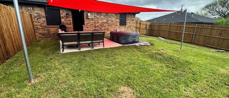 A backyard with a brick house, a wooden fence, a red shade sail, a bench, and a round bean bag.