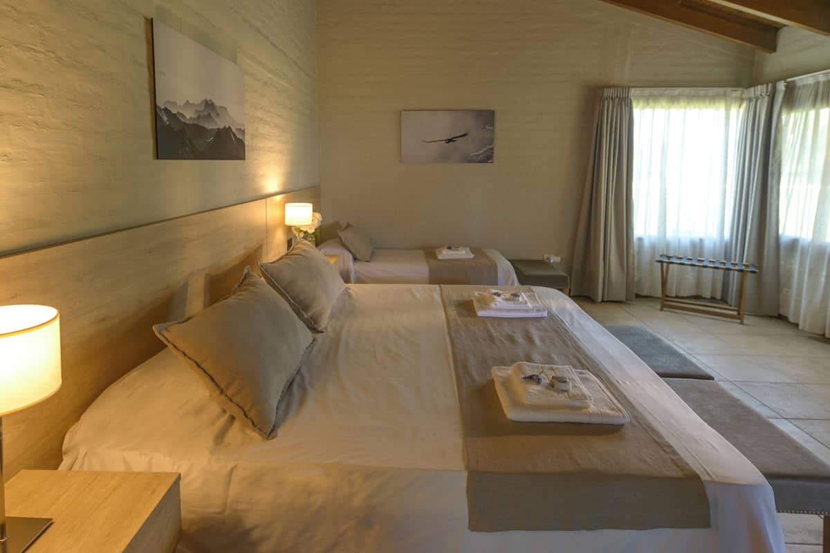Una habitación de hotel con una cama grande, otra más pequeña, una mesita de noche con lámpara, un reposabebidas con toallas y obras de arte en las paredes.