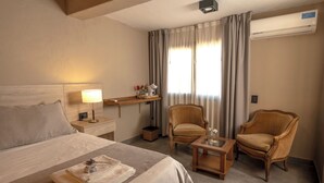 Una camera d'albergo con un letto, due poltrone, un tavolino, un comodino con una lampada, una scrivania con un computer e una finestra con tende.