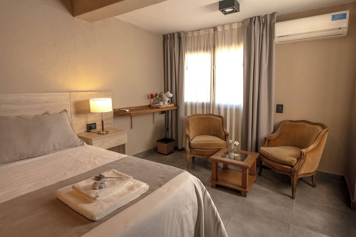 Habitación de hotel con cama, dos sillones, una mesita, una mesita de noche con lámpara, un escritorio con computadora y una ventana con cortinas.