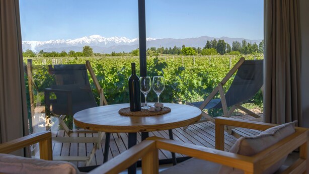 Un'area esterna con terrazza in legno, un tavolo e sedie, una bottiglia di vino e due bicchieri da vino. Si intravede una catena montuosa sullo sfondo.