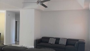 Un salon moderne avec un canapé et un pouf gris foncé, un ventilateur de plafond et une porte donnant sur une autre pièce.