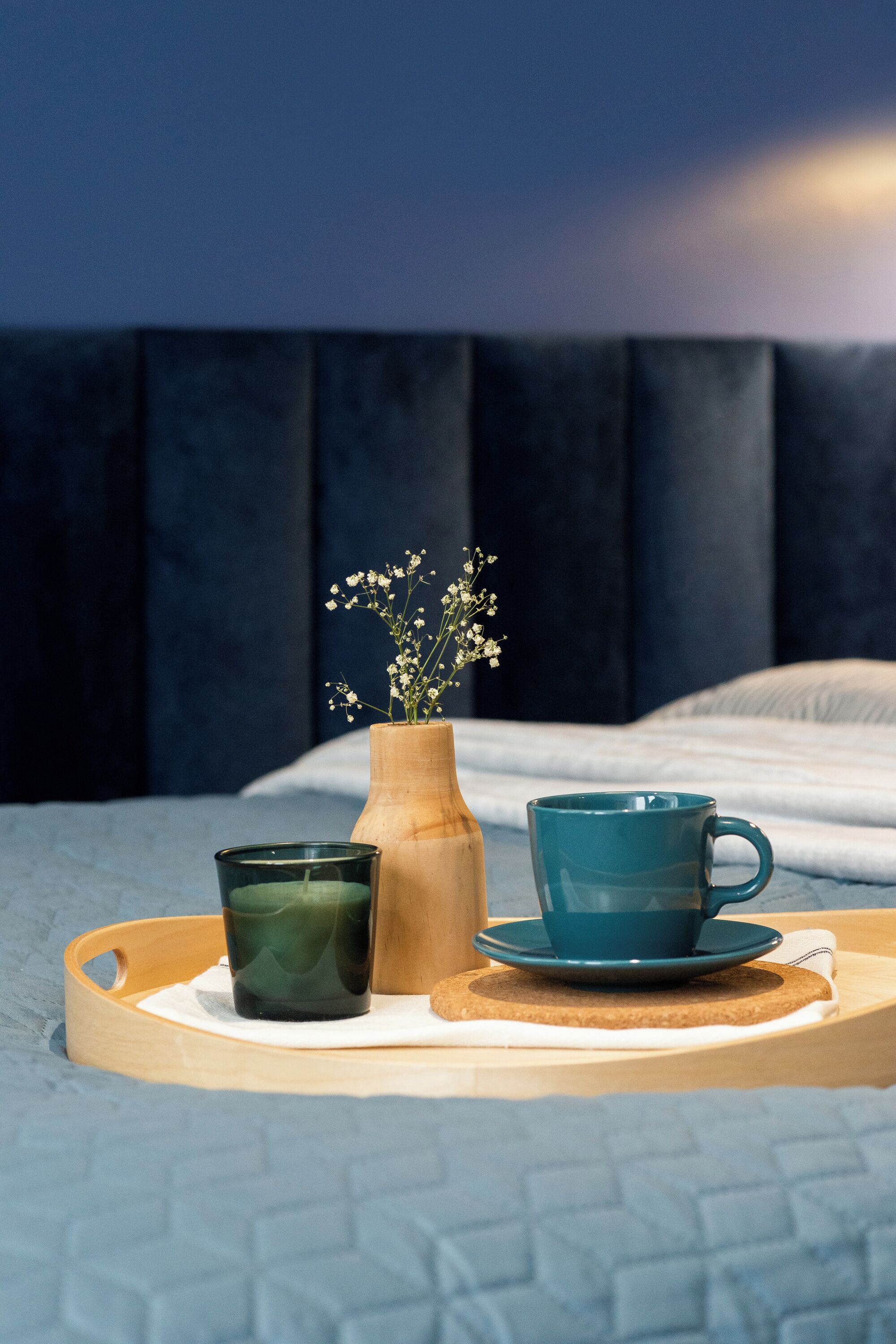 A bed with a dark headboard, a wooden tray with a green glass, a wooden vase with flowers, and a teal mug on a light-colored bedspread.