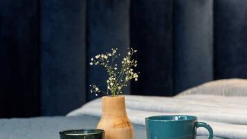 A bed with a dark headboard, a wooden tray with a green glass, a wooden vase with flowers, and a teal mug on a light-colored bedspread.