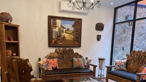 Una sala de estar con un sofá de madera, una mesa de centro y una araña. Hay un cuadro en la pared y una ventana con vistas a árboles.