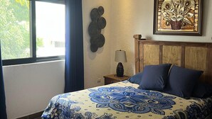 Un dormitorio con cama, ventilador de techo, ventana con cortinas, una obra de arte en la pared y una mesita de noche con lámpara.