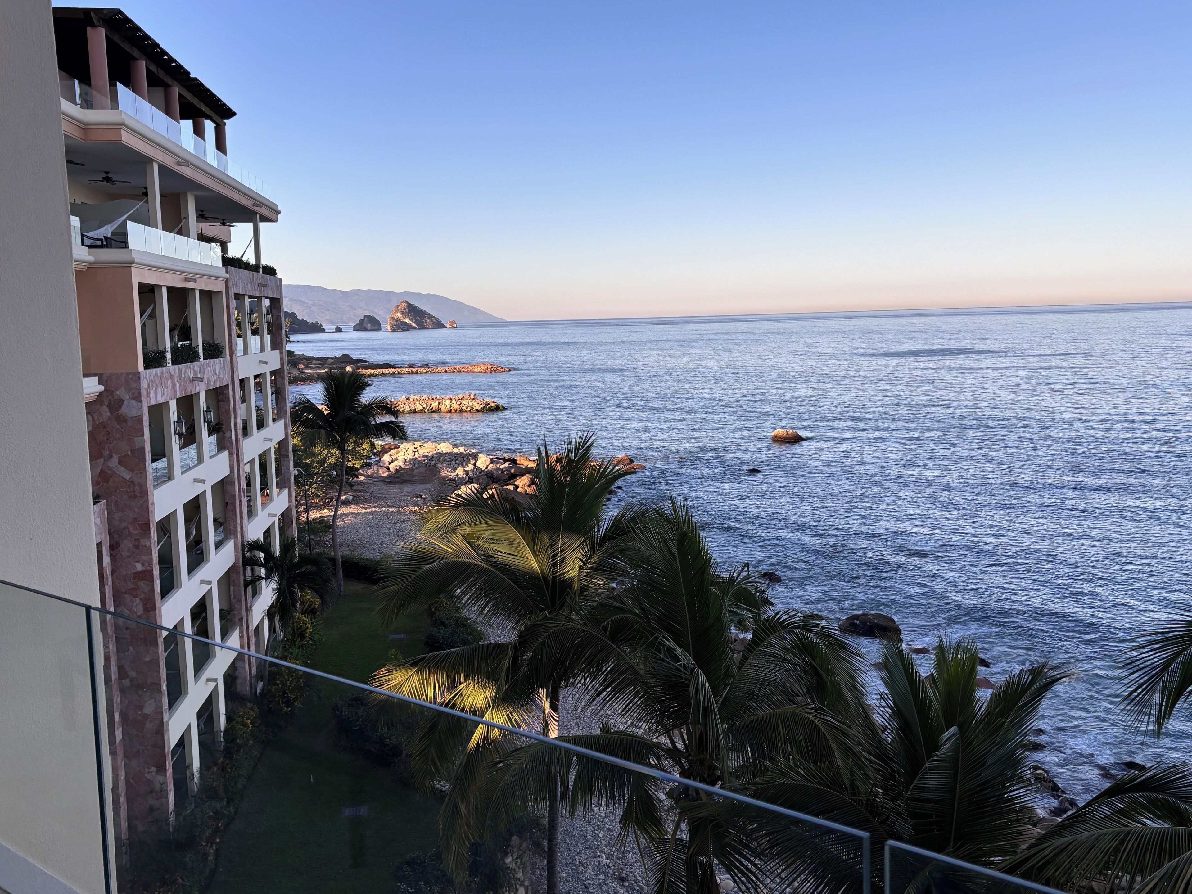 A coastal hotel with a balcony overlooking the sea, featuring a rocky shoreline and palm trees.