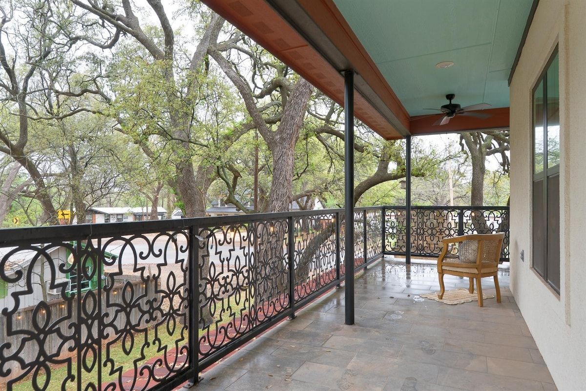 Een overdekt balkon met een decoratieve metalen balustrade, een houten stoel en uitzicht op bomen.