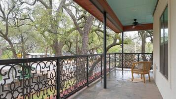 Een overdekt balkon met een decoratieve metalen balustrade, een houten stoel en uitzicht op bomen.