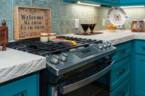 A kitchen with a gas stove, oven, and teal cabinets. There is a sign that reads 'Welcome Mi casa Es su casa' and a decorative plate on the counter.
