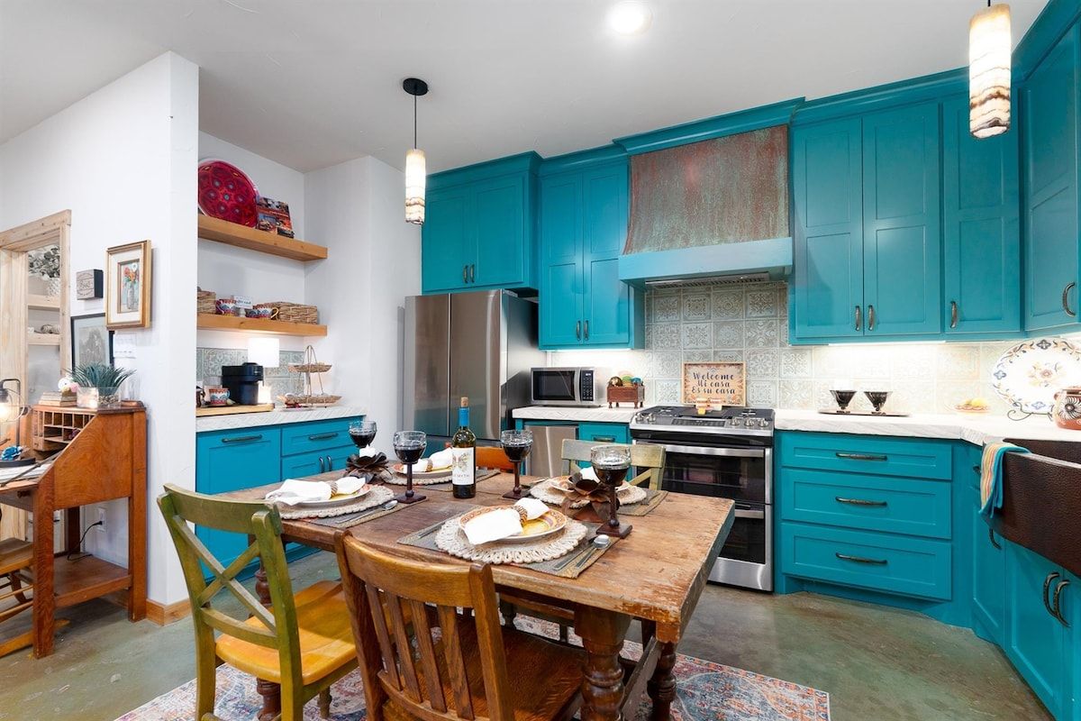 Een keuken met blauwgroene kastjes, een houten eettafel en diverse keukenapparaten.