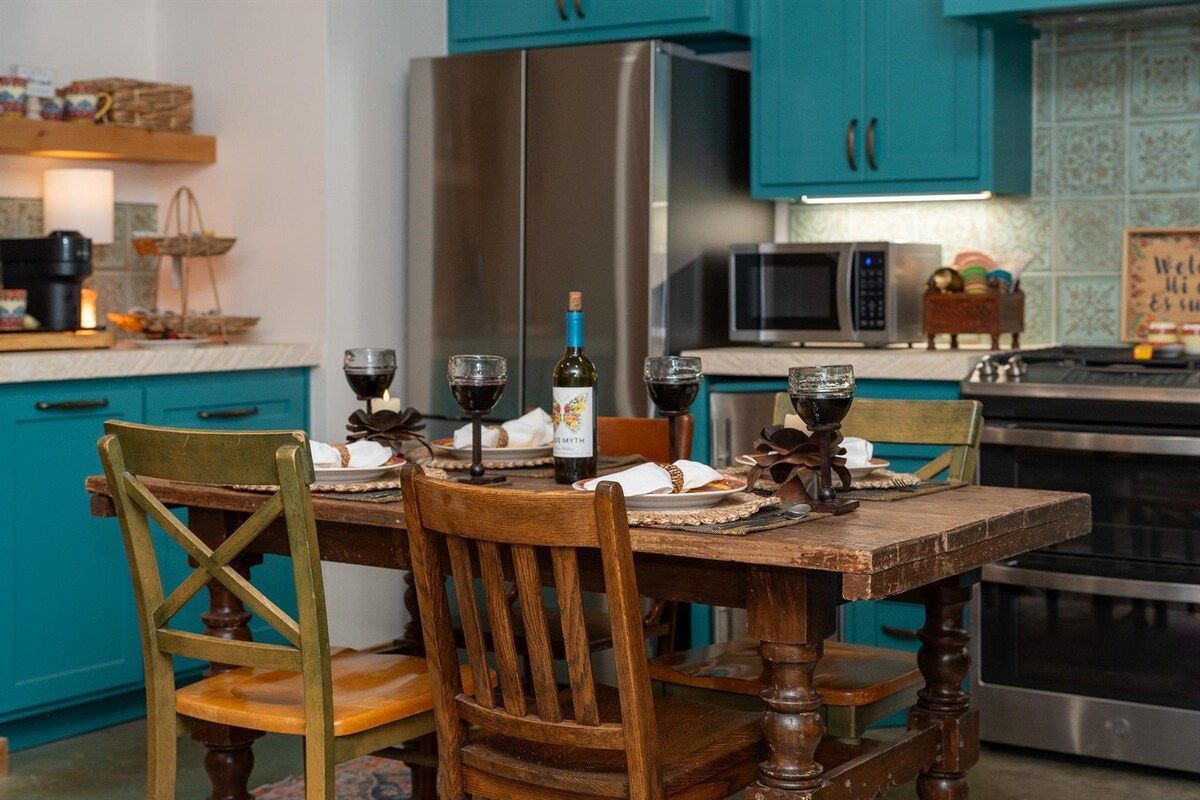 Une cuisine avec des meubles de couleur turquoise, un réfrigérateur en acier inoxydable, un four à micro-ondes et une table à manger agrémentée de verres à vin et d’une bouteille.