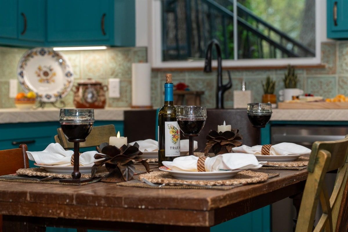 A dining table set with wine glasses, a bottle, and a plate in a kitchen with teal cabinets and a window.