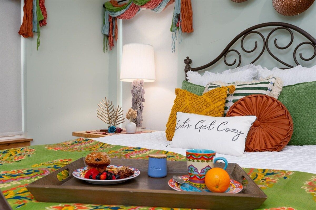 Een net opgemaakt bed met een kussen waarop staat 'Let's Get Cozy', een dienblad met gebak en fruit, en een lamp op een nachtkastje.