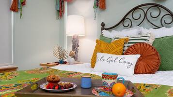 Een net opgemaakt bed met een kussen waarop staat 'Let's Get Cozy', een dienblad met gebak en fruit, en een lamp op een nachtkastje.